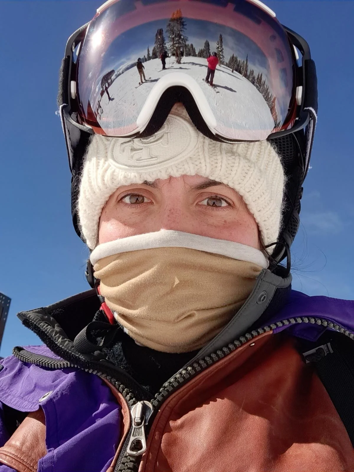 A person wearing ski gear, including a helmet with goggles, a white knitted hat, a face covering, and a jacket, taking a selfie outdoors on a snowy mountain with other skiers and snow-covered trees reflected in the goggles under a clear blue sky.