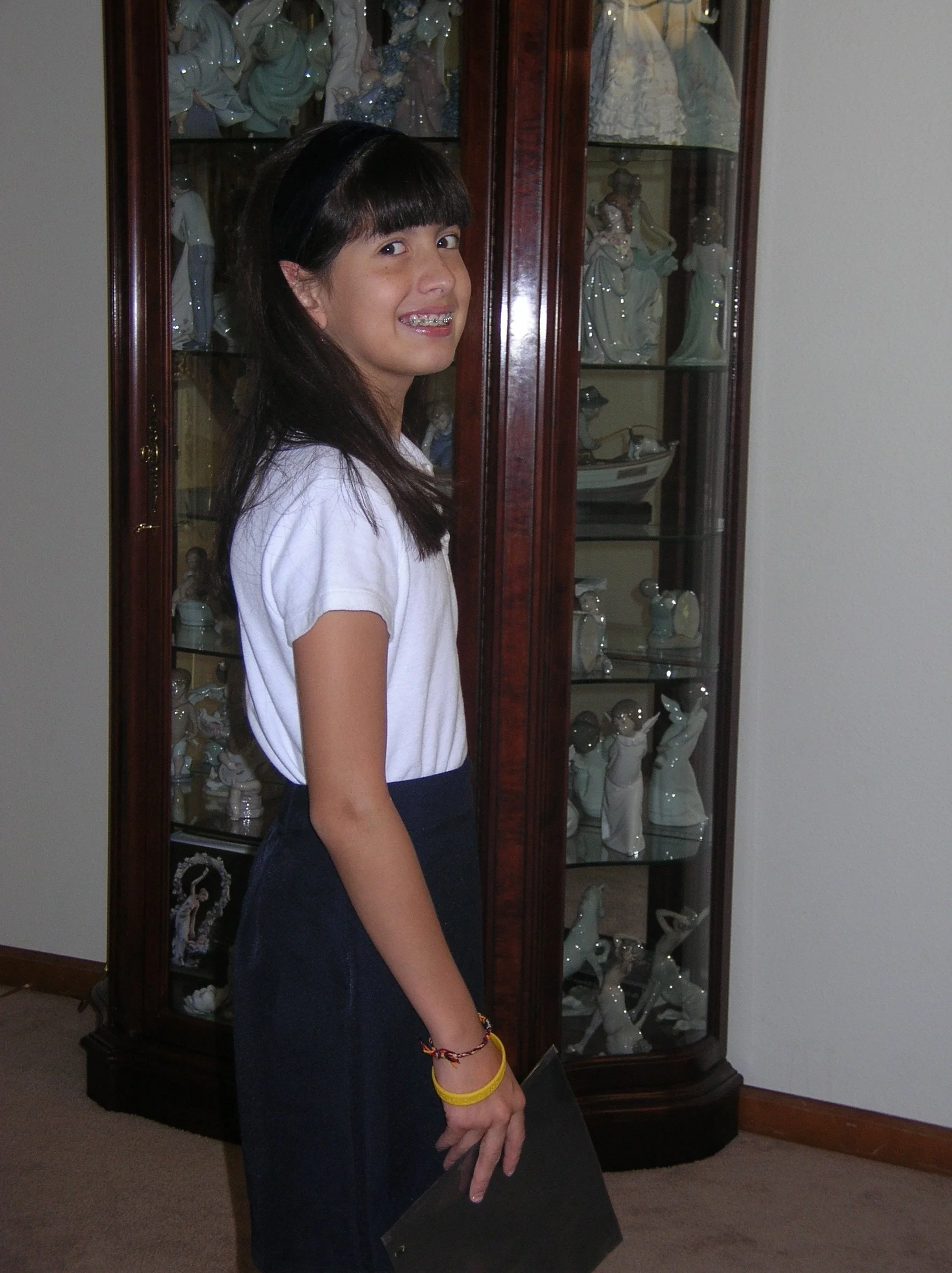 A girl with long dark hair and a big smile, wearing a white shirt and a dark skirt, stands indoors in front of a glass display cabinet filled with porcelain figurines, including angels and other decorative items.