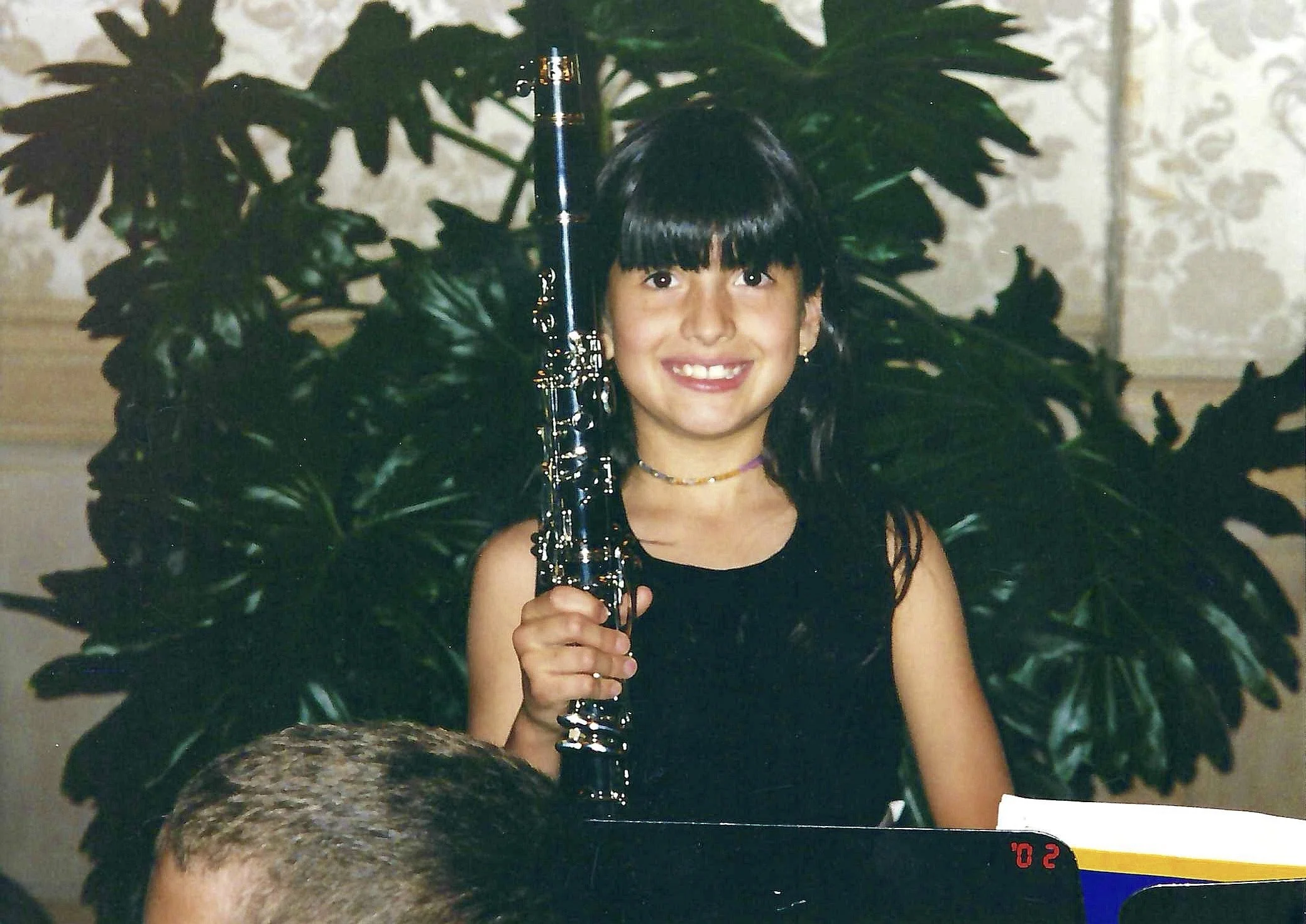A young girl with dark hair and bangs, smiling, holding a glass of champagne, surrounded by large green leaves in an indoor setting.