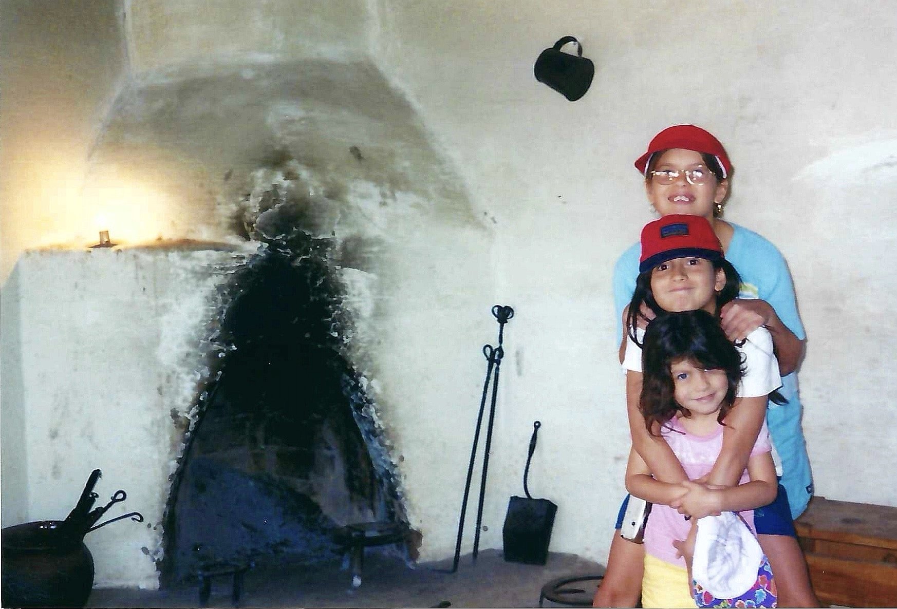 Three children, two girls and a boy, are standing together, smiling and hugging. They are indoors with a plain white wall behind them. The girl in the middle has glasses and a red cap, the girl on the front is wearing a pink top and yellow shorts, an