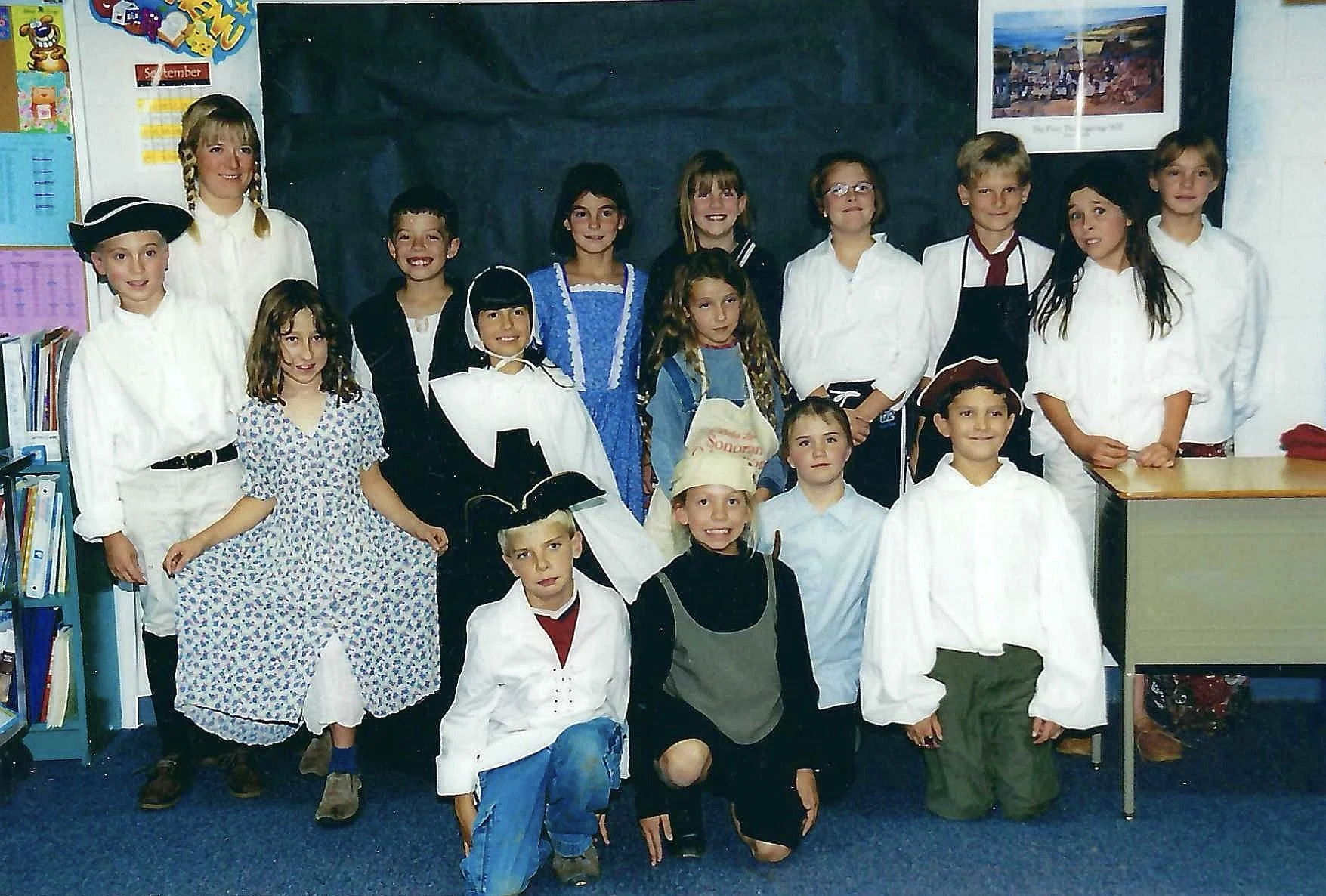 Group of children in historical costumes posing in a classroom with bookshelves and a blackboard.