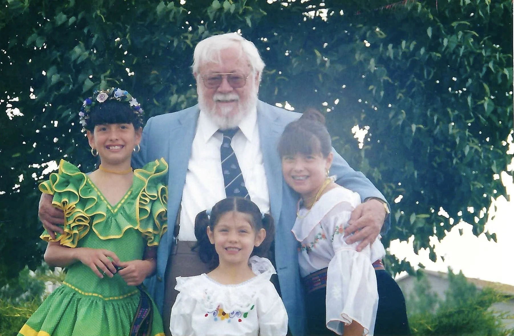 An elderly man with white hair and beard wearing glasses, a blue suit, and a tie, standing outdoors in front of a green leafy background, surrounded by four young girls with dark hair, wearing colorful traditional dresses, smiling for the photo.