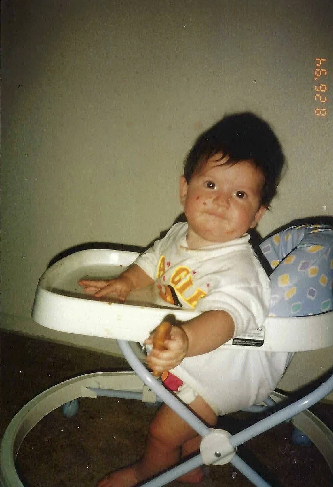 A young child sitting in a high chair with mess on their face and hands, smiling and reaching out with a messy hand.