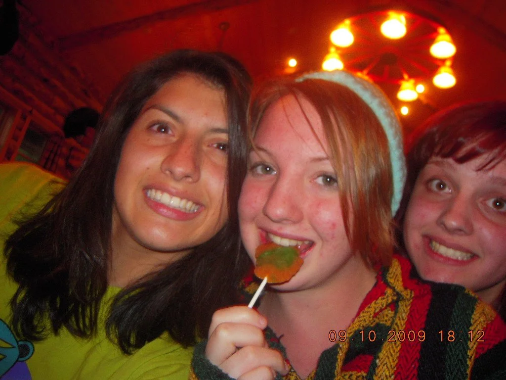 Three friends smiling at a party, one holding a colorful leaf-shaped candy on a stick, with warm, orange lighting in the background.