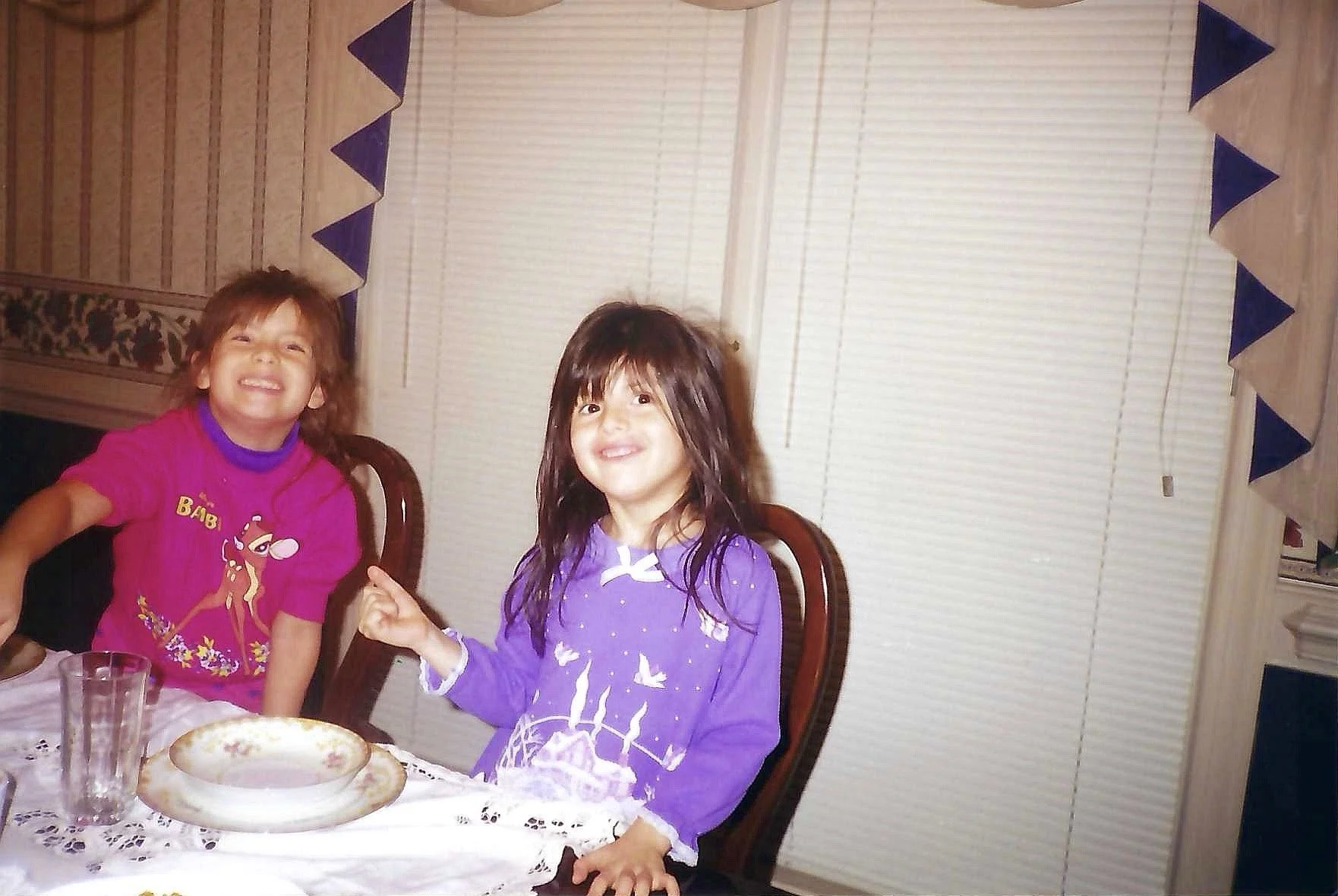 Two young girls at a birthday celebration sitting at a table with a cake and glasses, smiling and pointing, in a room with blinds and blue valance curtains.