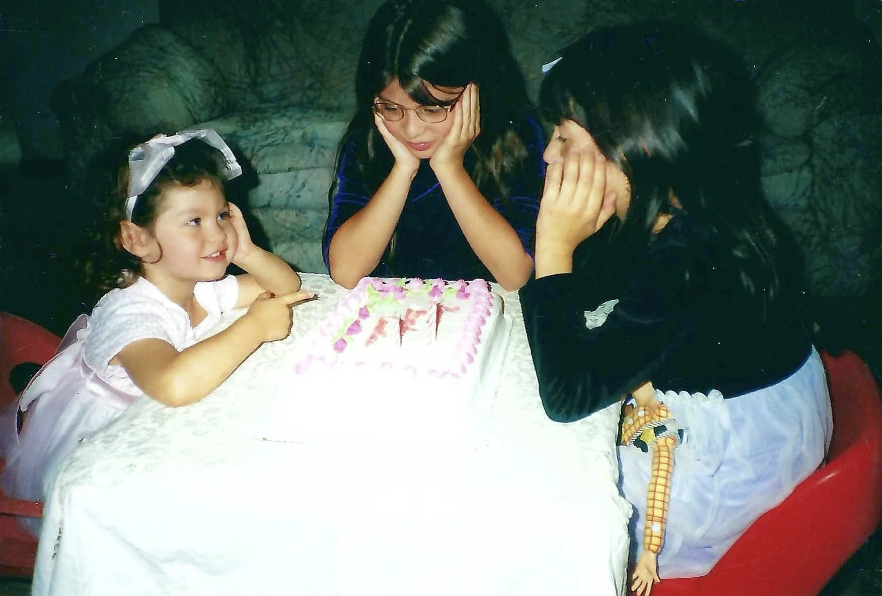 Three females, a young girl and two women, are gathered around a birthday cake on a table. The girl is wearing a pink dress with a large bow in her hair and is pointing at the cake. The women, one with glasses and long hair and the other with dark ha