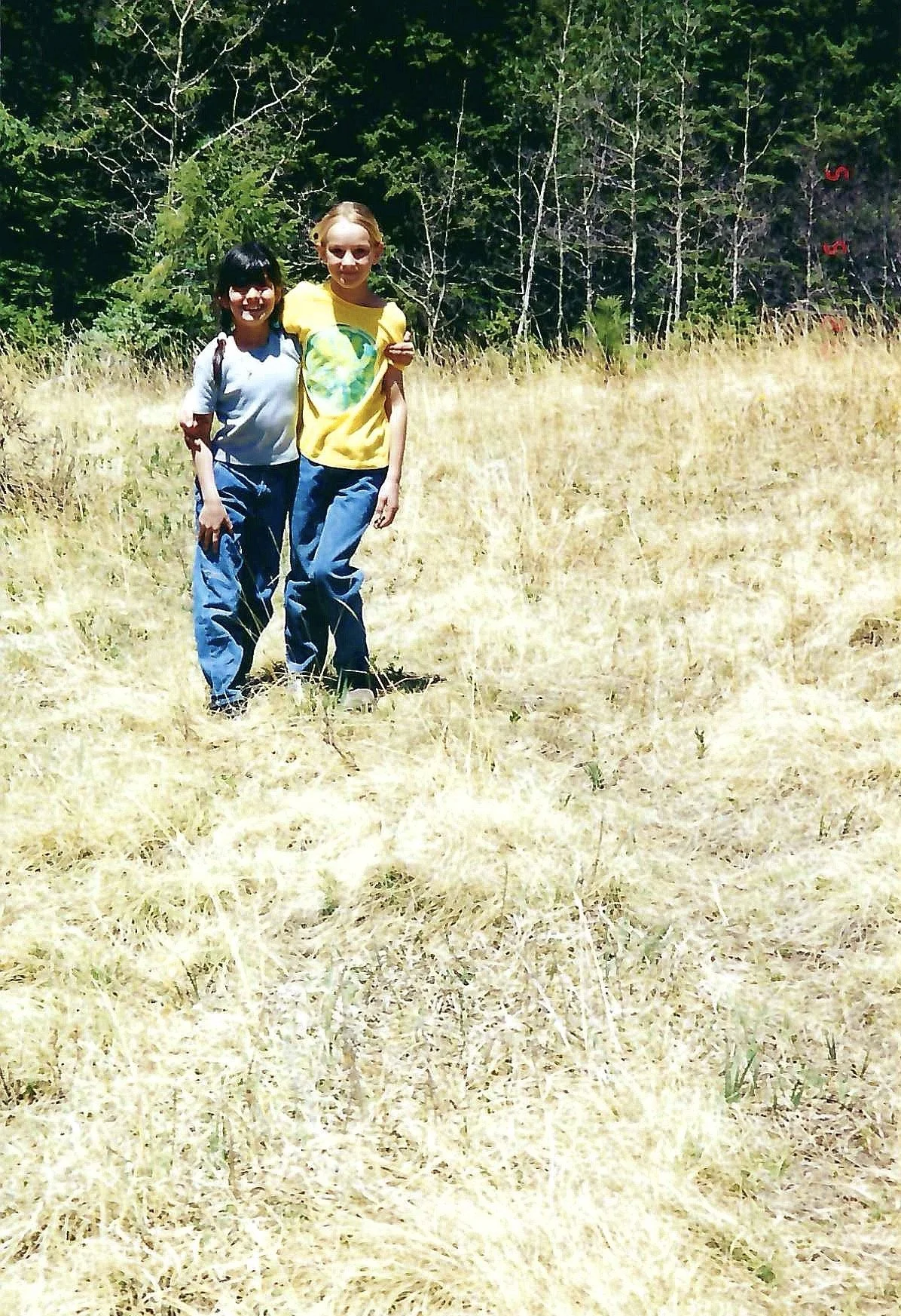 Two young girls standing outdoors in a grassy field, with trees in the background, smiling at the camera.