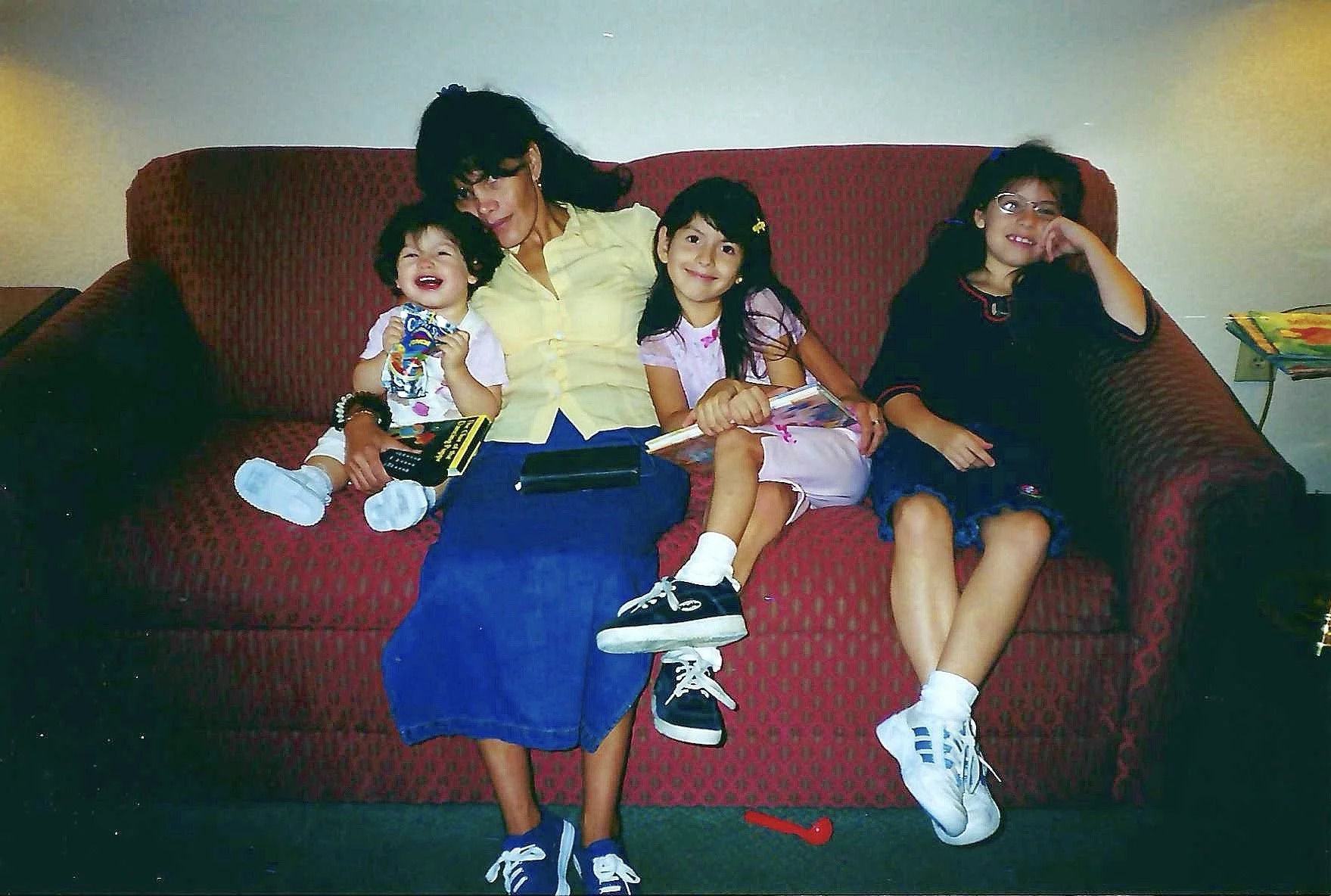 Four children and one adult sitting on a red patterned couch in a living room, with some books and toys around.