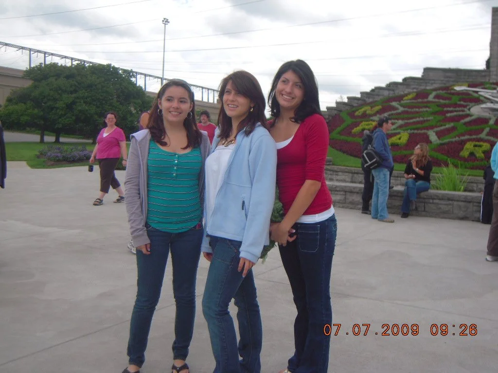 Three young women standing together outdoors, smiling, with a flower bed and other people in the background.