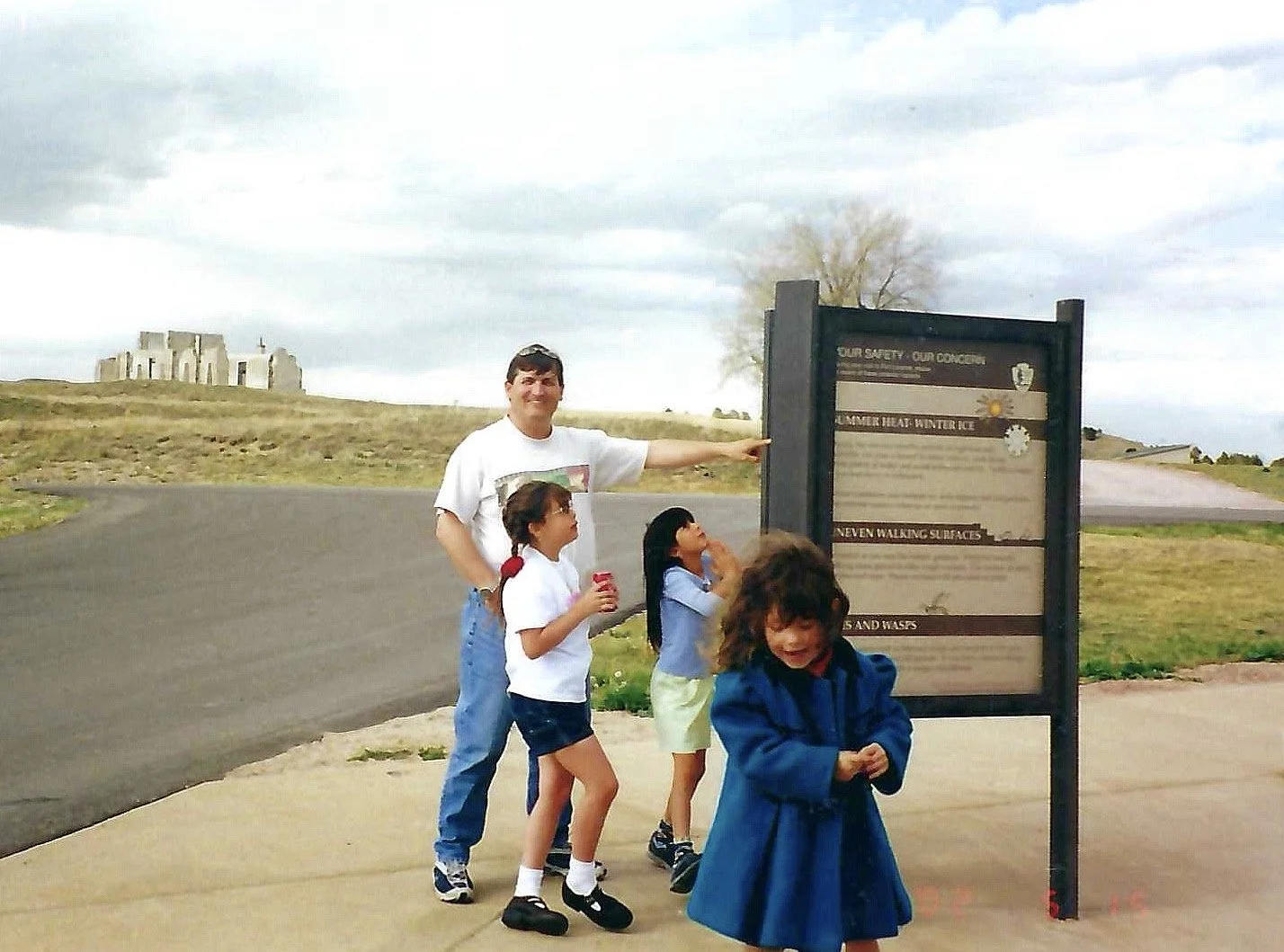A group of four children and an adult standing outdoors near a signboard at a historical site, with a grassy hill and ruins in the background on cloudy day.