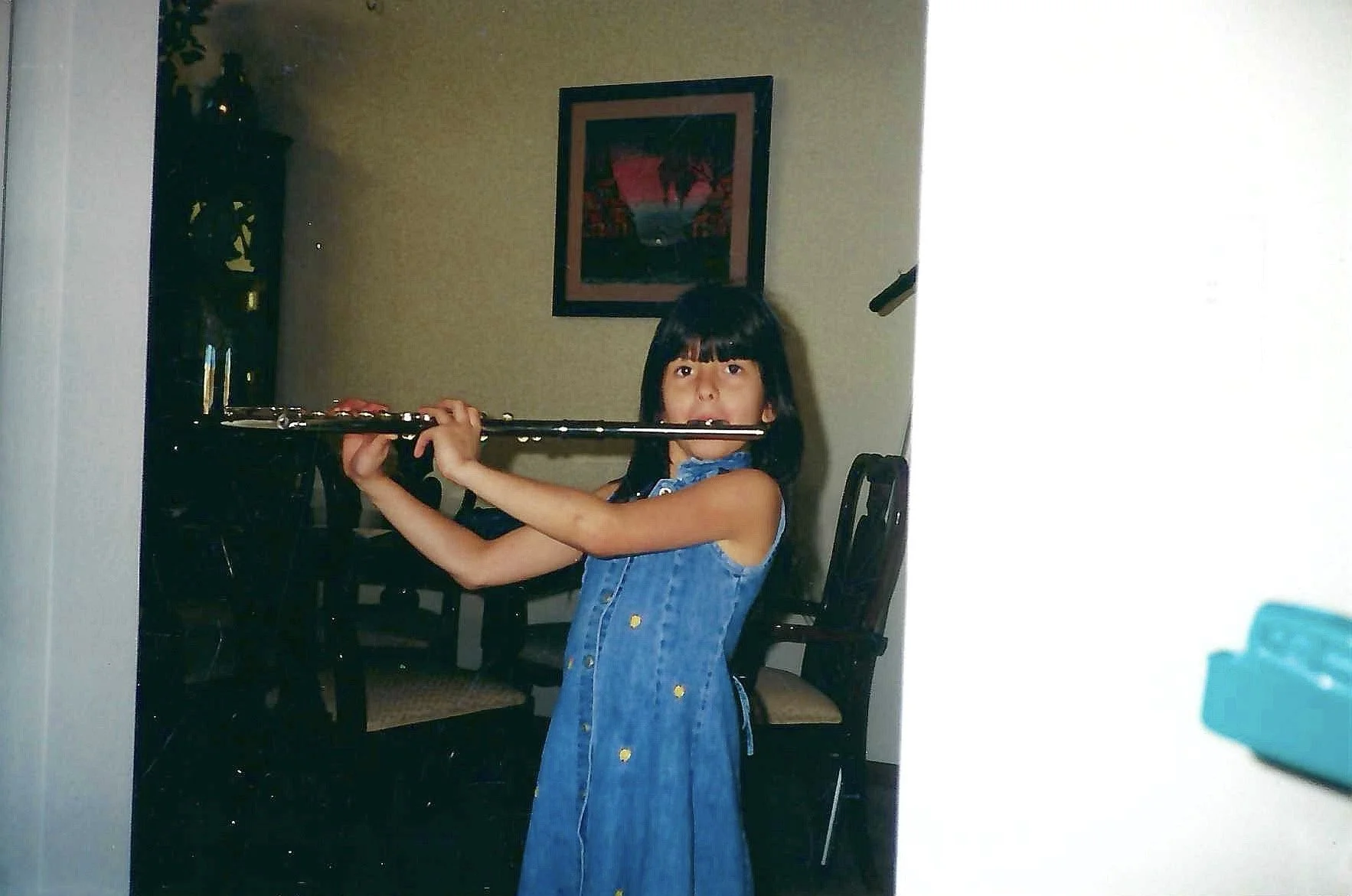 A young girl with dark hair and bangs, wearing a blue denim dress with buttons, is holding a flute across her chest in a dining room with a painting on the wall and chairs around the table.
