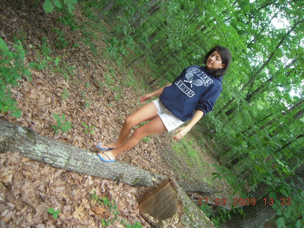 A young woman standing on a fallen tree trunk in a forest, surrounded by green trees and leaves. She is wearing a dark hoodie, white shorts, and flip-flops. The timestamp on the photo indicates it was taken on July 3, 2009 at 1:22 PM.