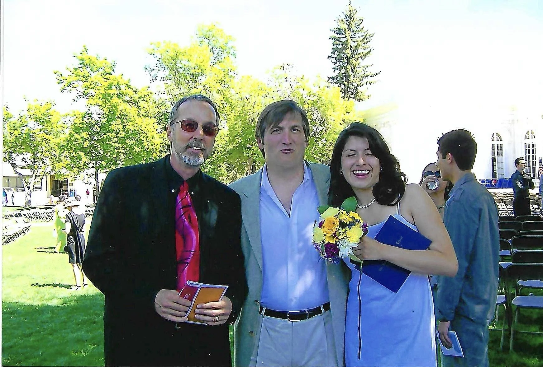 Three people standing outdoors, celebrating a graduation, with chairs and people in the background.