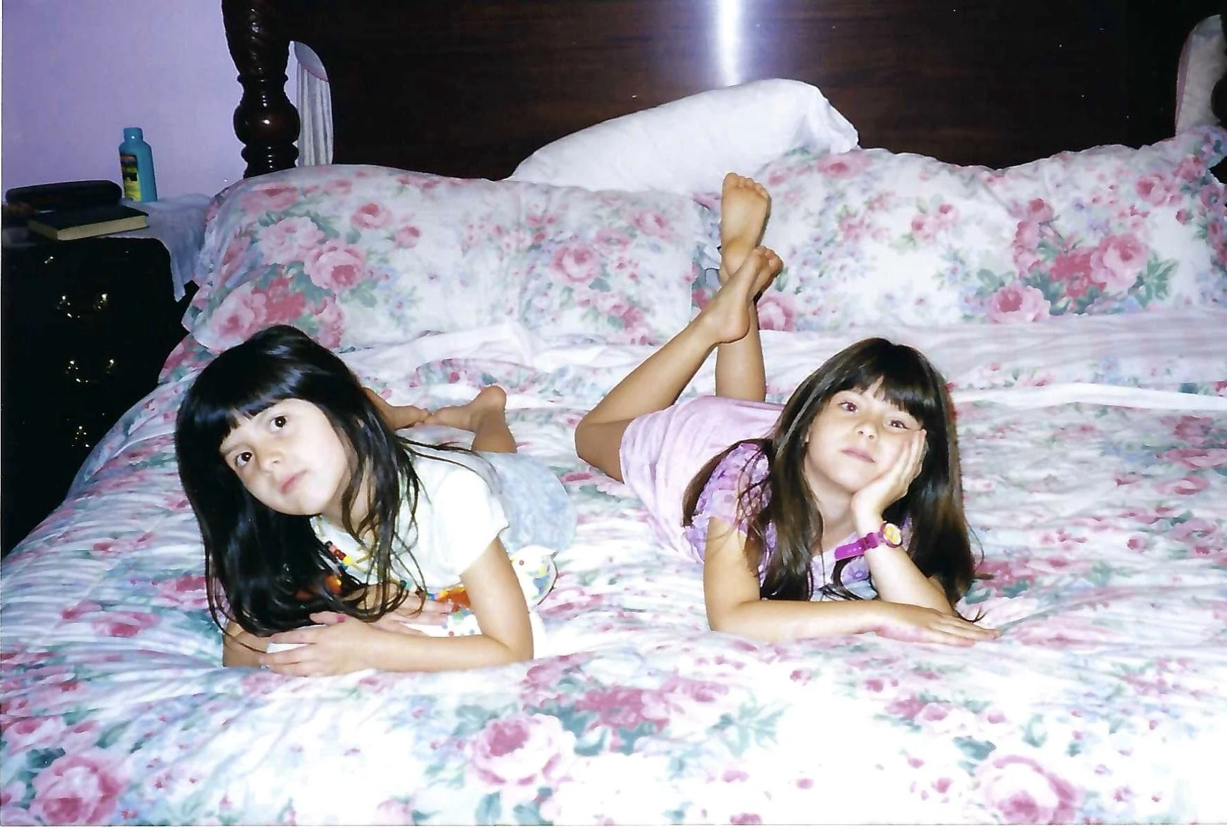 Two young girls with dark hair are lying on a floral bedspread with pink and green patterns, resting on their elbows with hands under their chins, looking at the camera.