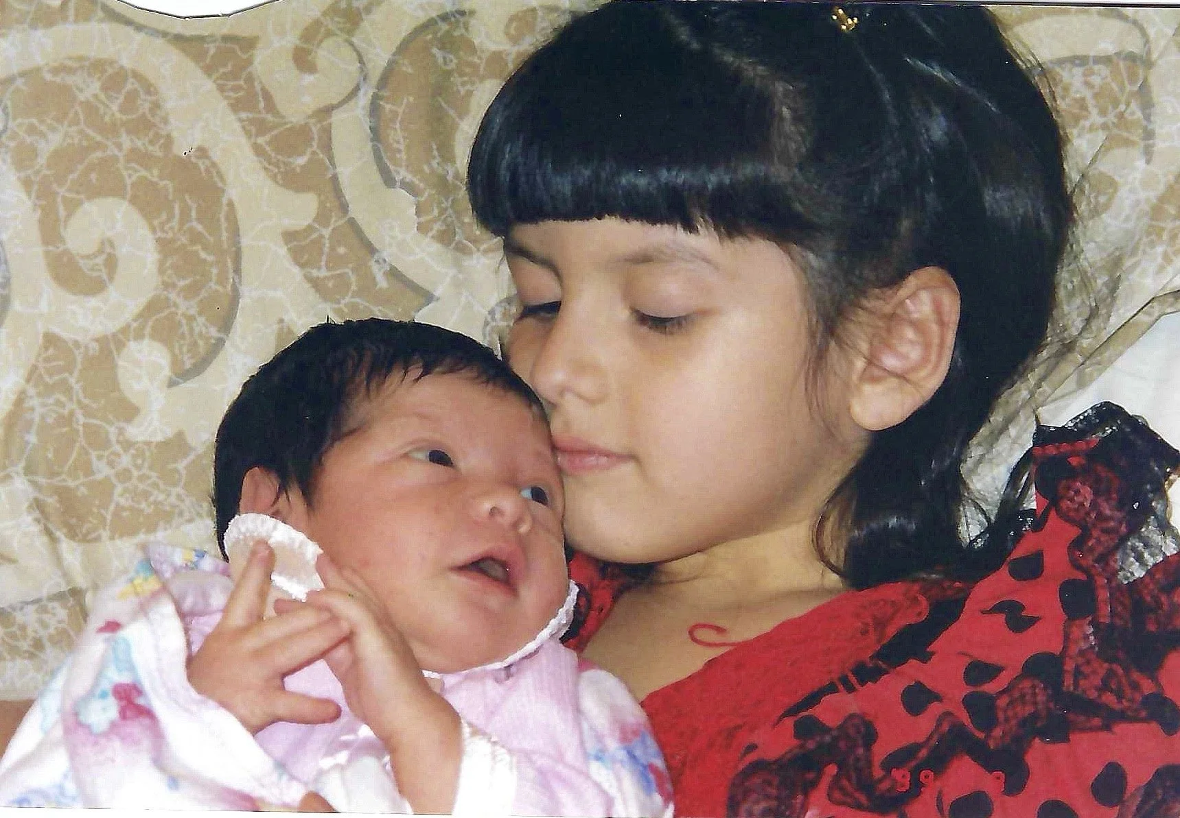 A young girl and a newborn baby sharing a tender moment. The girl is holding the baby close, and they are touching foreheads with eyes closed.