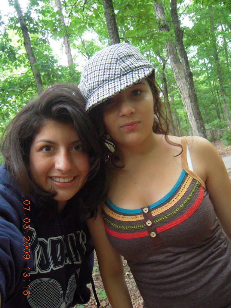 Two young women taking a selfie outdoors in a forest with green trees visible in the background.