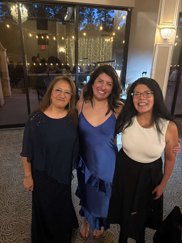 Three women smiling and posing together inside a decorated event space, with fairy lights and a glass door behind them.