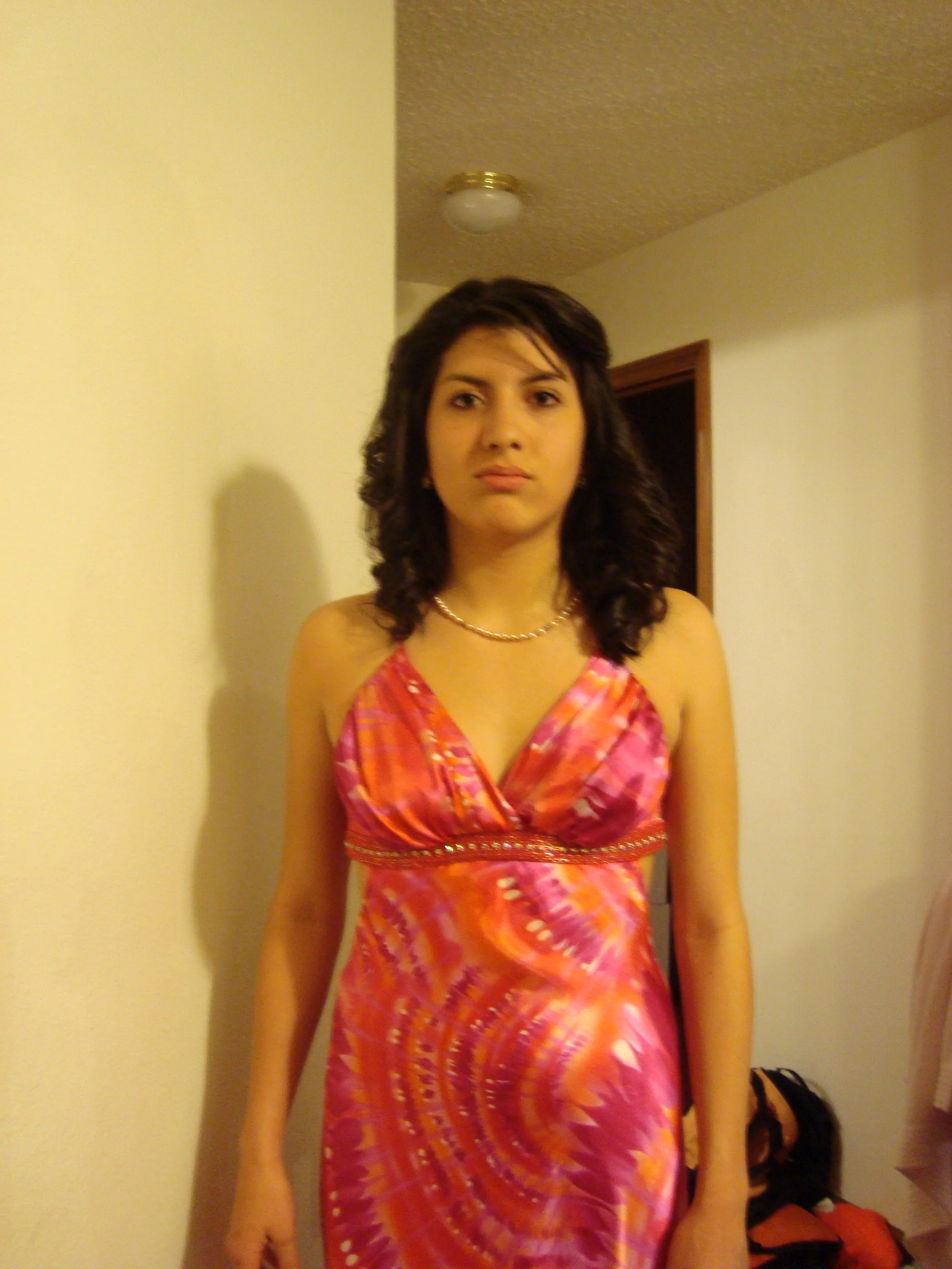 A woman standing inside a room wearing a colorful pink, orange, and purple dress with spaghetti straps, a pearl necklace, and shoulder-length wavy dark hair. The room has plain light-colored walls, a ceiling light fixture, and some bags or clothing o