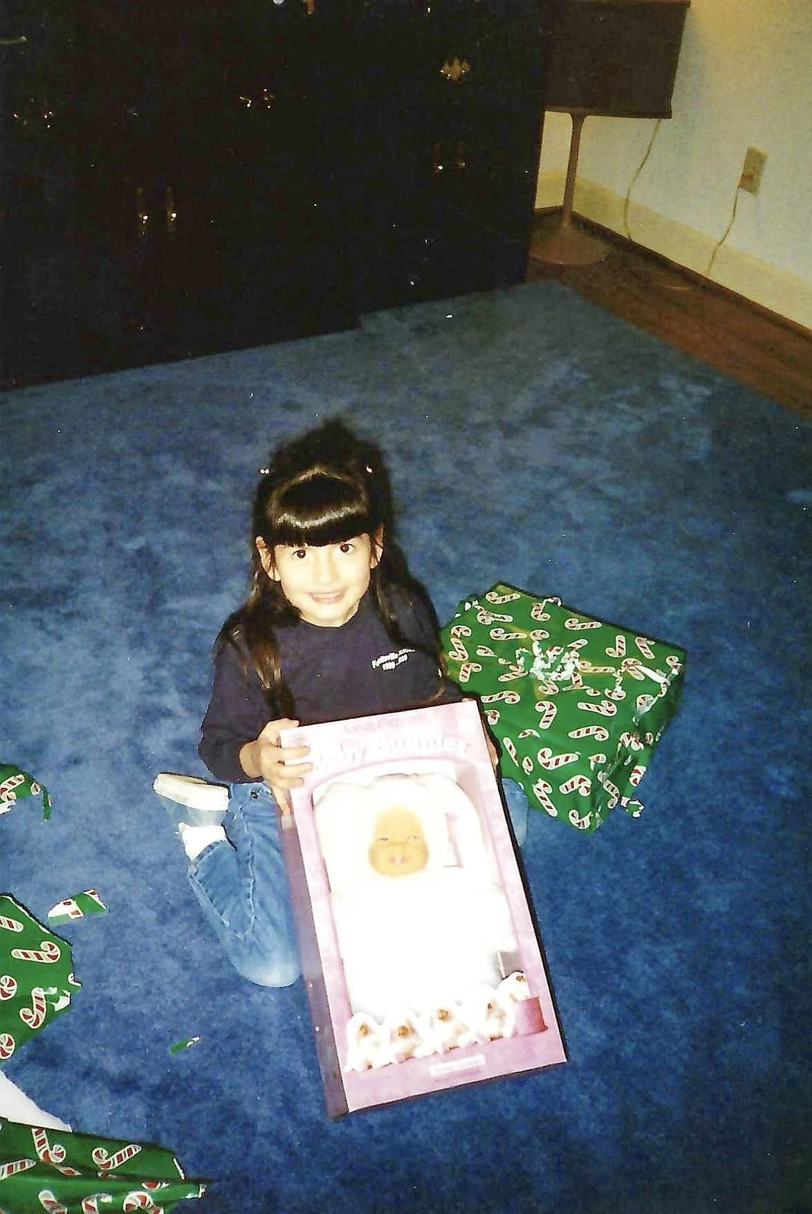 A young girl with dark hair and a bow in her hair, sitting on a blue carpeted floor, holding a pink box with a picture of a baby on it, surrounded by wrapped Christmas presents, with a dark dresser and a small table in the background.