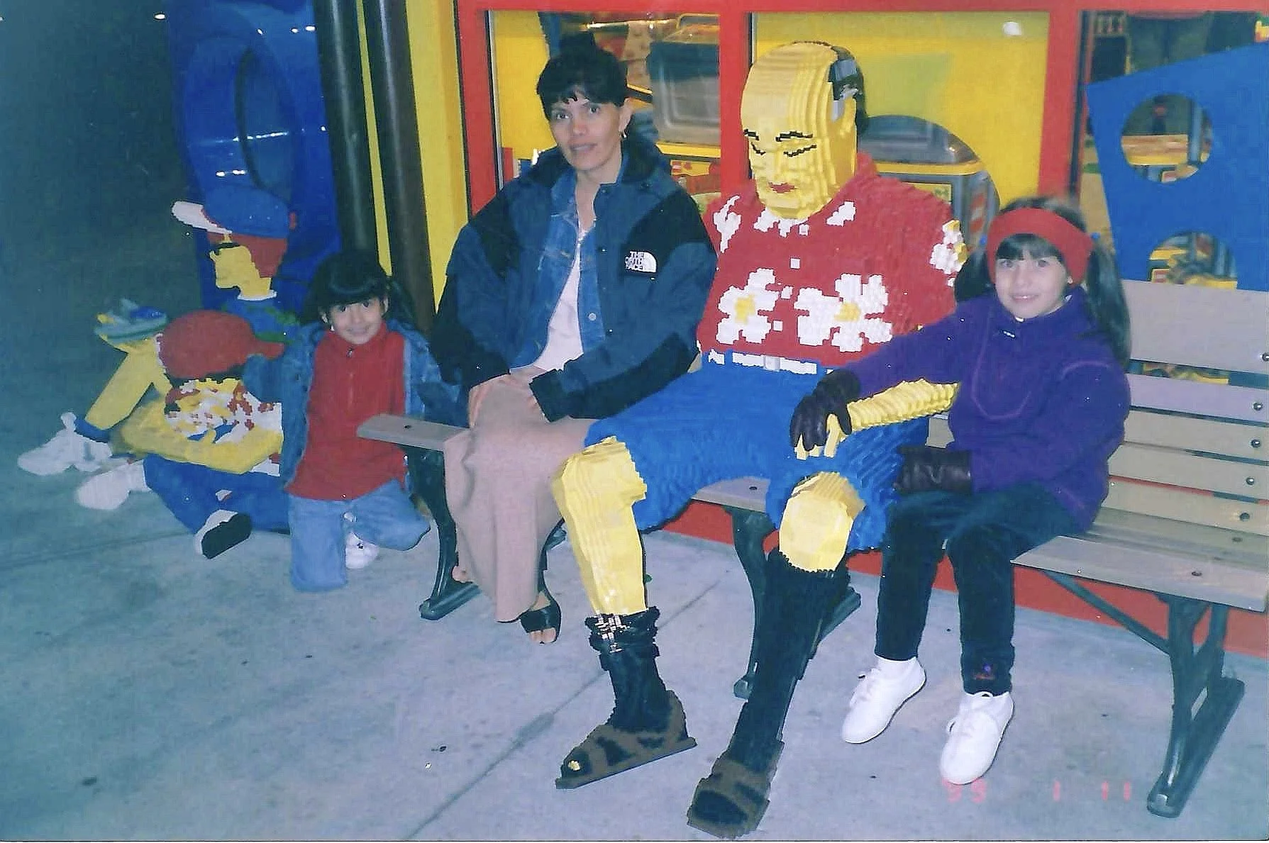 Three children sitting on a park bench with two life-sized LEGO sculptures of a woman in the middle and a man on the right. The children are dressed in warm clothing, and the LEGO sculptures are built with yellow, red, and blue LEGO bricks.