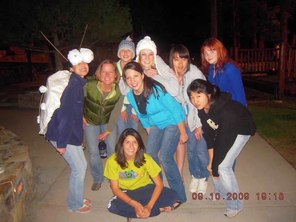 Group of nine young women smiling and posing for a photo outdoors at night, with some wearing colorful jackets and beanies, standing and sitting on a concrete path with a wooden fence and trees in the background.