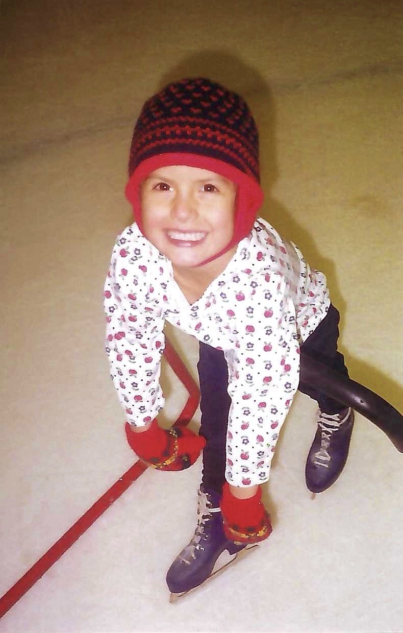 A young child in winter clothing, wearing a colorful knit hat, a patterned white jacket with small red and black designs, black pants, red gloves, and ice skates, sitting on an ice rink near the edge.