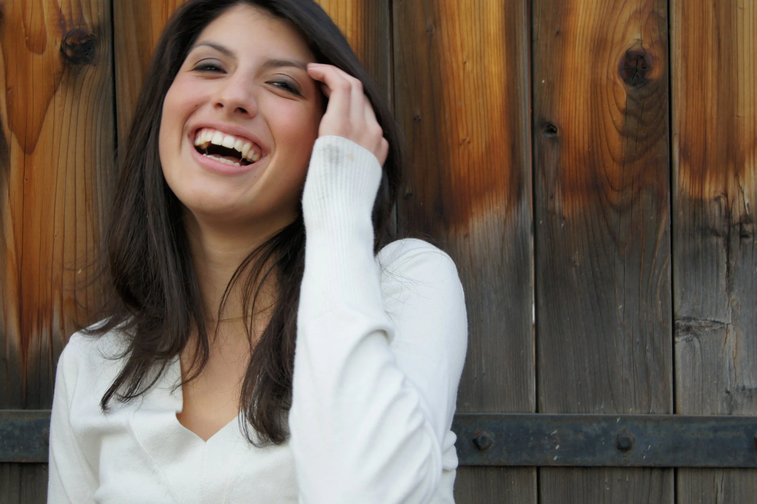 A woman with long dark hair laughing, wearing a white sweater, standing in front of a wooden fence.