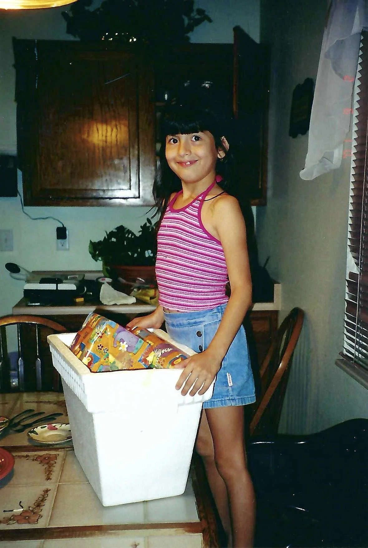 A young girl with long dark hair wearing a pink striped tank top and a denim skirt, standing in a kitchen, holding a colorful gift-wrapped box. There are cabinets, a window with blinds, and various kitchen items in the background.