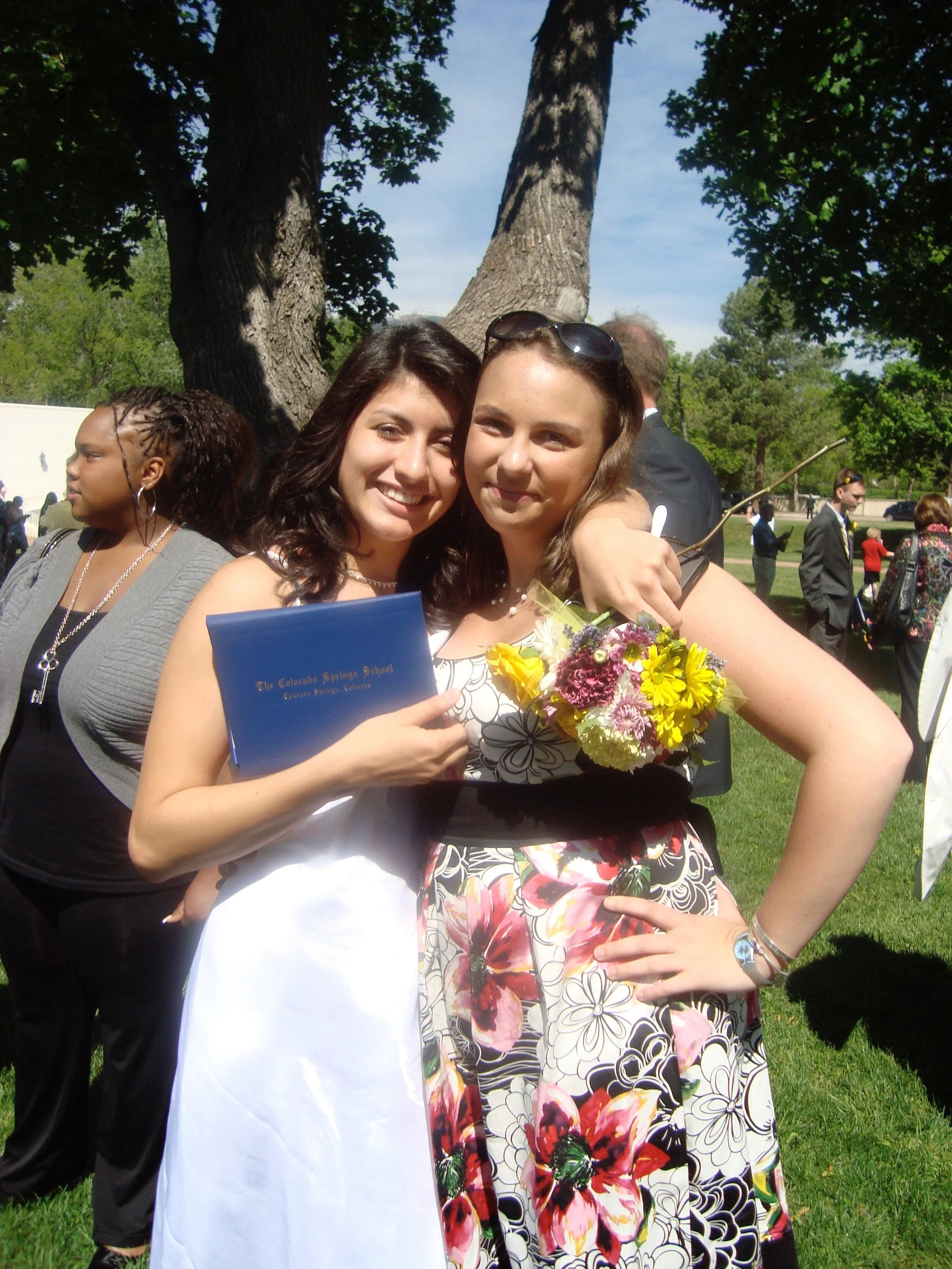 Two young women are hugging outside during what appears to be a graduation celebration. The woman on the left is smiling and holding a diploma from The Colorado Springs School, while the woman on the right is holding a bouquet of flowers and has her 