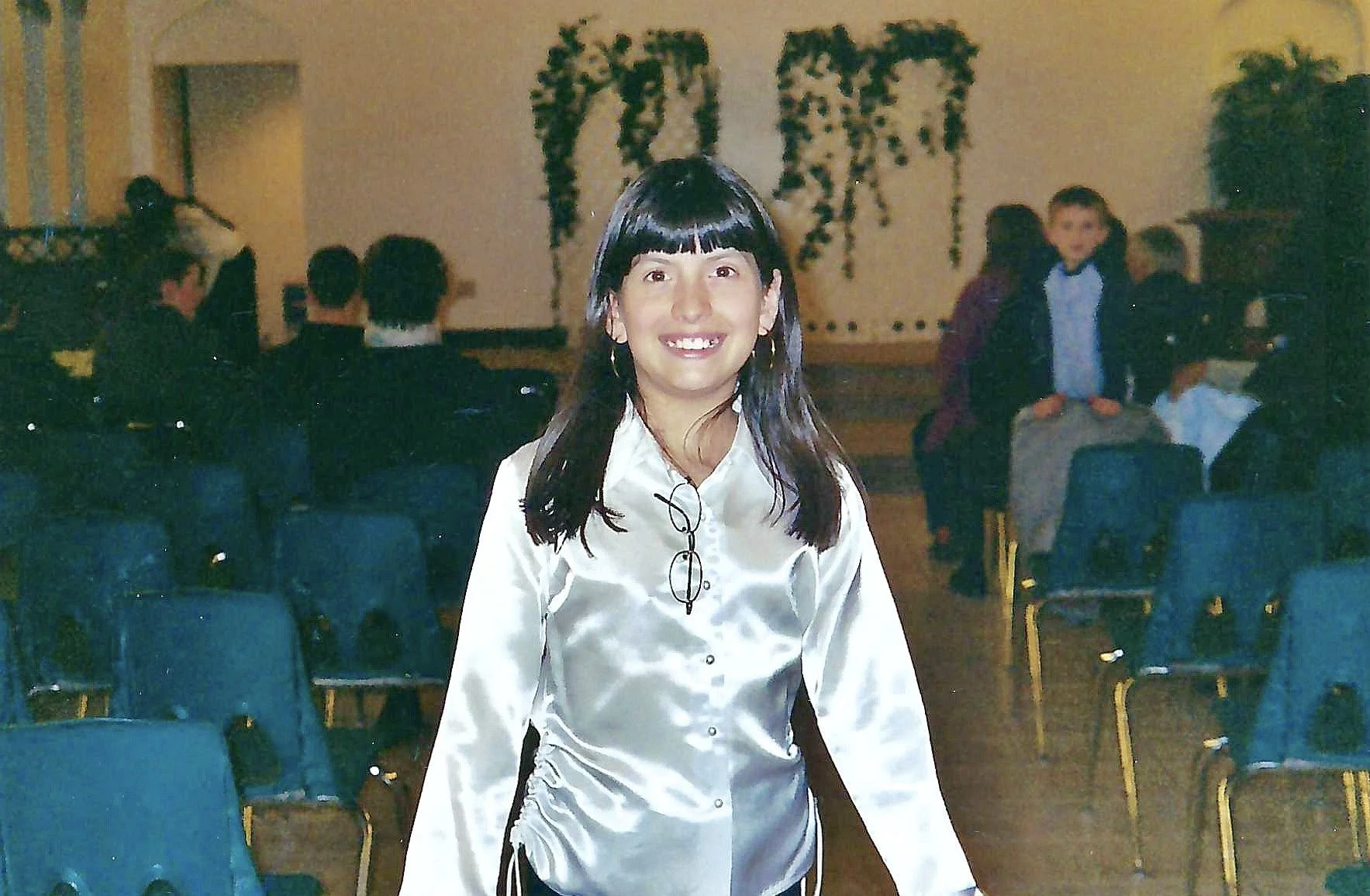 A woman with black hair, glasses hanging from her shirt, wearing a white satin blouse, smiling at the camera in a decorated indoor space with people talking and seated in background.