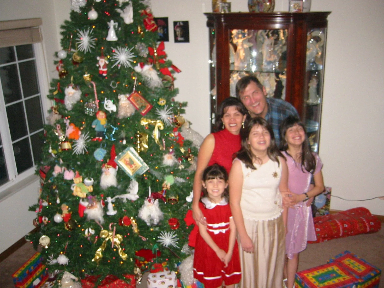 A group of five people, including children and an adult man, standing and smiling next to a decorated Christmas tree with wrapped presents underneath. The room has a window on the left and a dark wooden display cabinet on the right.