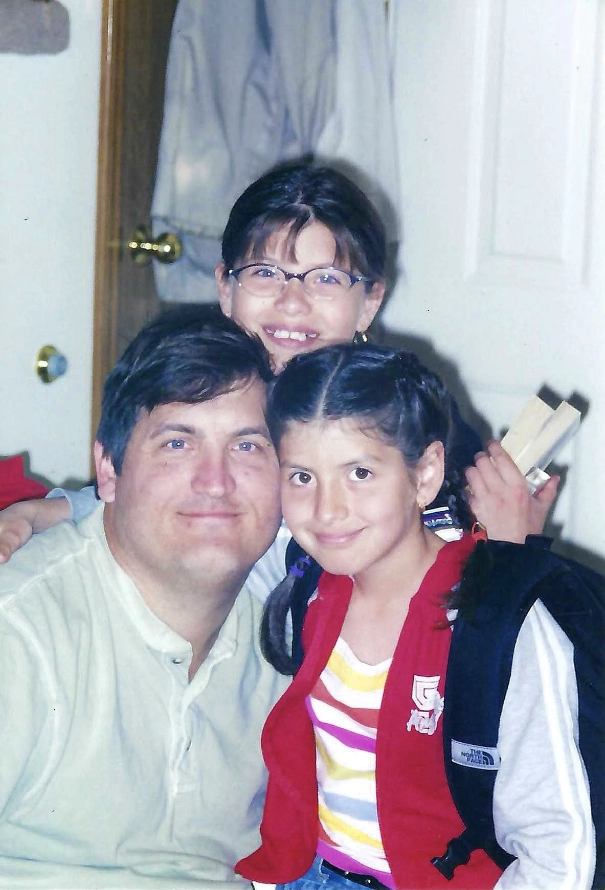 A man, a young girl, and a woman smiling for a photo indoors. The man is on the left, wearing a light-colored shirt. The young girl is in front, wearing a red jacket and a striped shirt, with braided hair and a backpack. The woman is behind them, wit