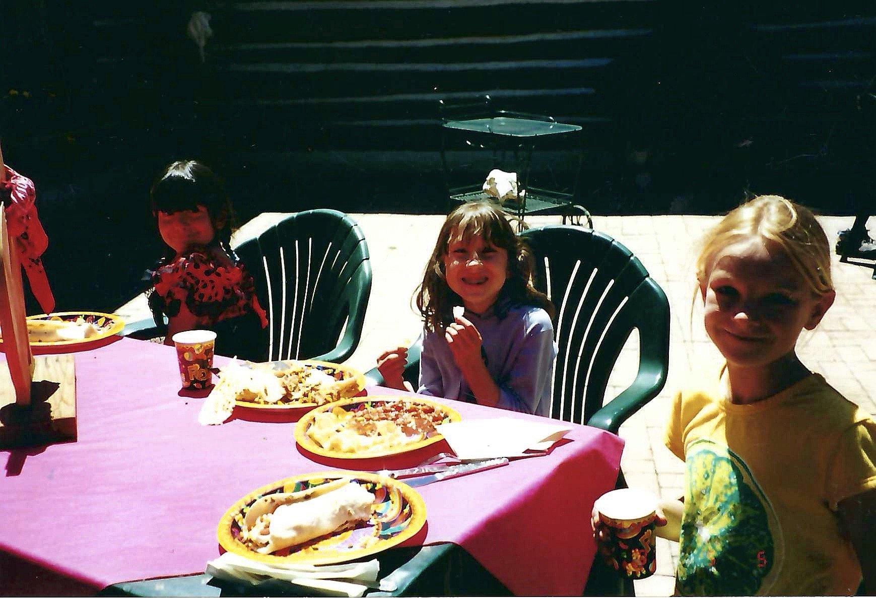 Children sitting at a table outdoors celebrating, with pizza and snacks on pink tablecloth, smiling and enjoying a party.