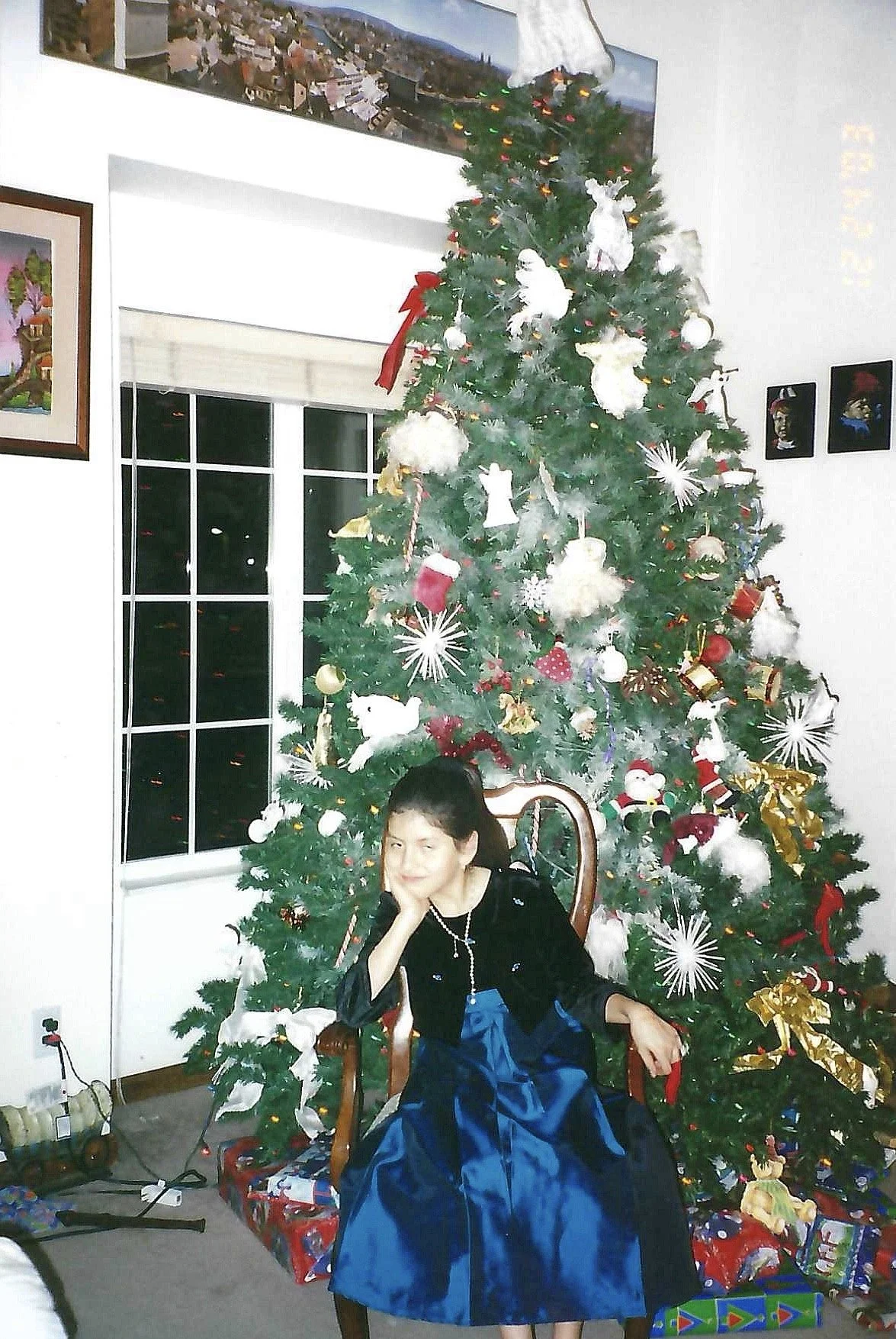 A woman sitting on a wooden chair in front of a decorated Christmas tree with ornaments and gifts, inside a room with a window, artwork on the walls, and a view of a snowy night outside.