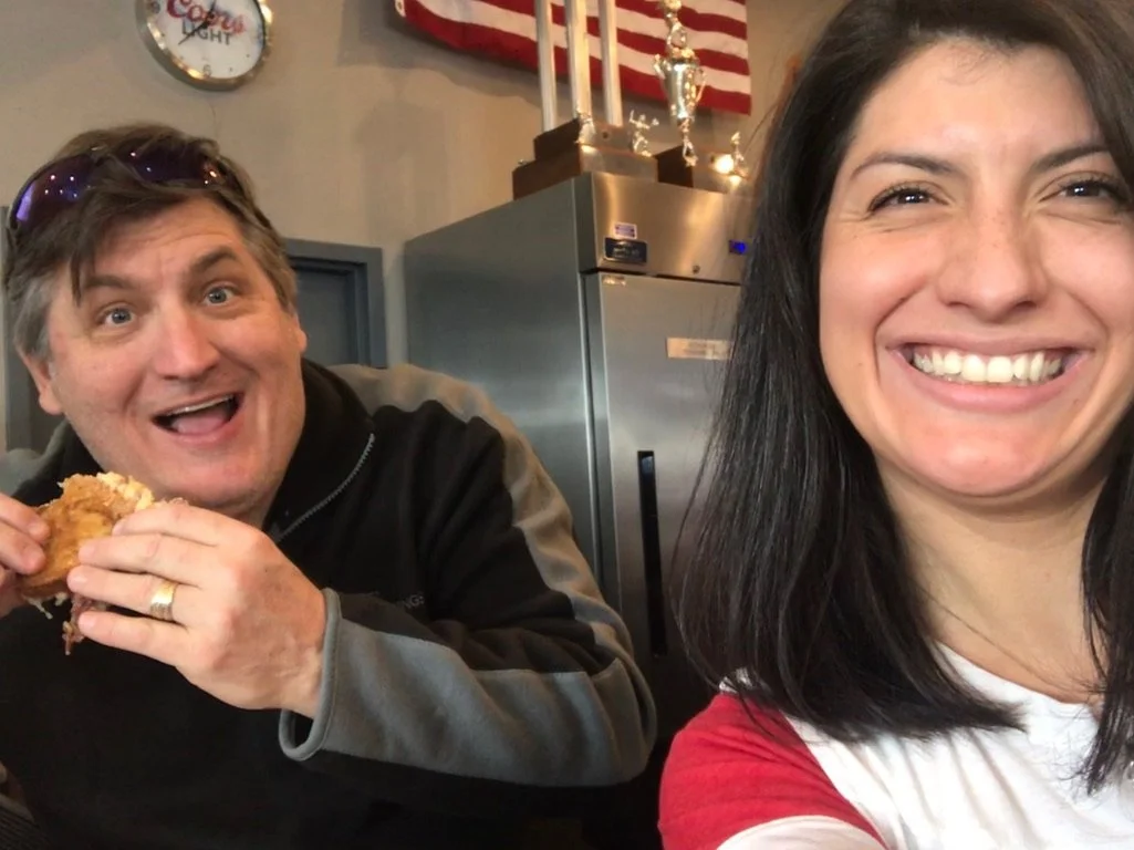 A man and woman smiling at a restaurant, with the man holding a piece of fried chicken, American flag and a wall clock in the background.