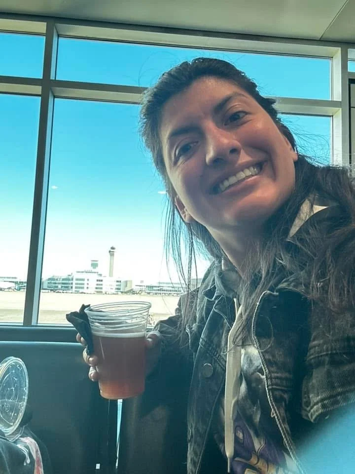 A woman smiling and holding a clear plastic cup with a beverage inside, sitting at an airport terminal with large windows and a view of the airport runway and control tower outside.