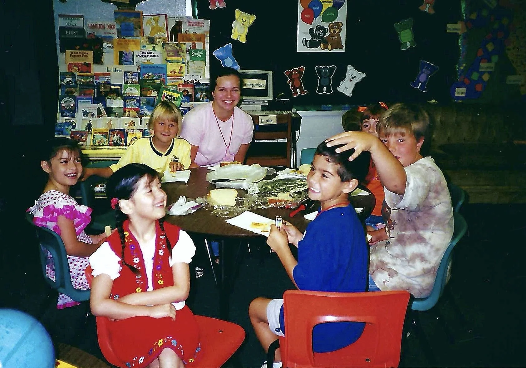 Young children and a teacher gathered around a table in a classroom, celebrating a birthday with cake.