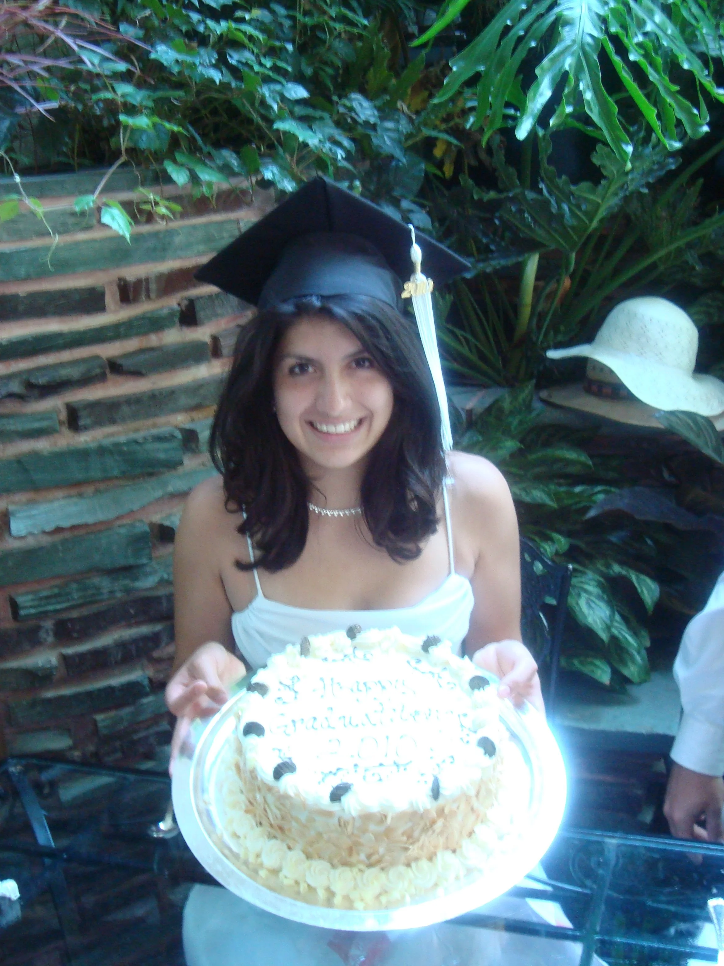 A woman with long dark hair wearing a white dress and a graduation cap with a tassel, smiling and holding a large birthday cake decorated with cream and writing, standing in front of green plants and a brick wall.