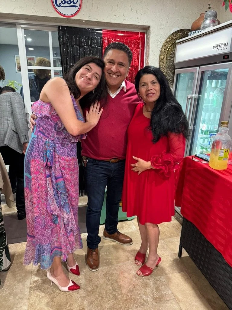 Three people standing together at a celebration, smiling and posing for a photo, with a decorated background and a refrigerator nearby.