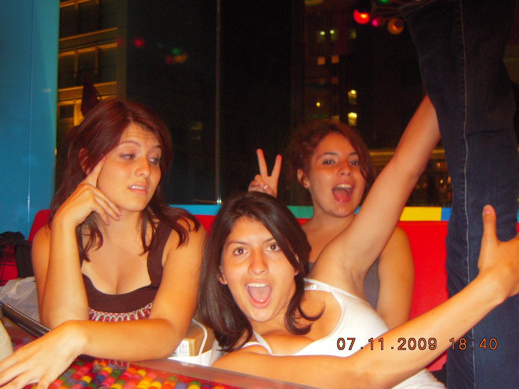 Three young women sitting at a table, two making peace signs, one posing with a surprised expression, with colorful background and a person standing nearby.