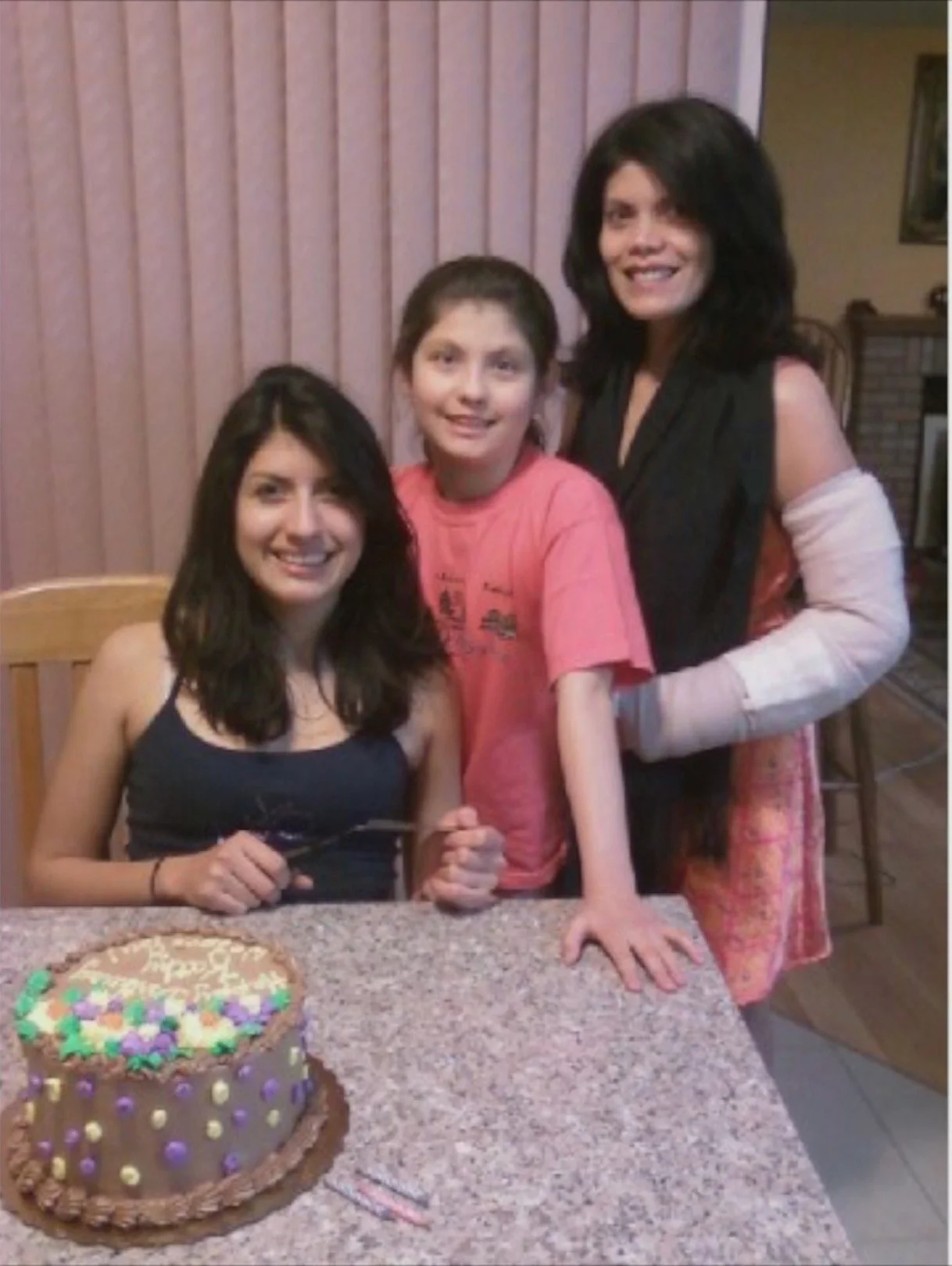 Three females at a birthday celebration are posing behind a decorated birthday cake on a granite countertop. The woman on the left is seated with dark hair and a sleeveless black top, the boy in the middle is standing in a pink shirt, and the woman o