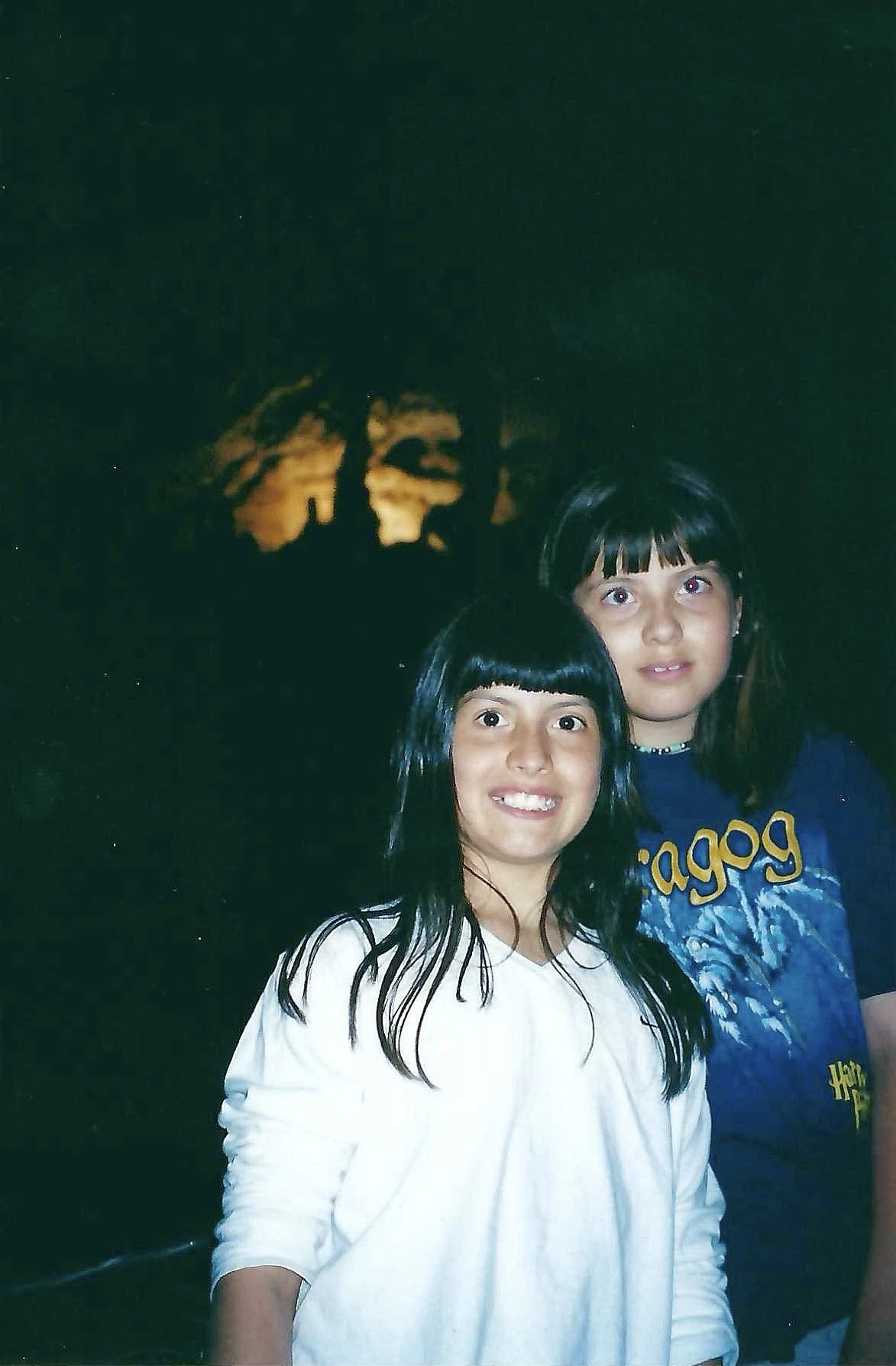 Two young girls with dark hair and bangs stand close together, smiling at the camera, in front of a dark background with a faint illuminated silhouette of trees or clouds.
