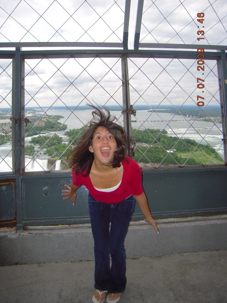 Woman in a red shirt standing on a high tower platform with a safety net and view of a river and city in the background.