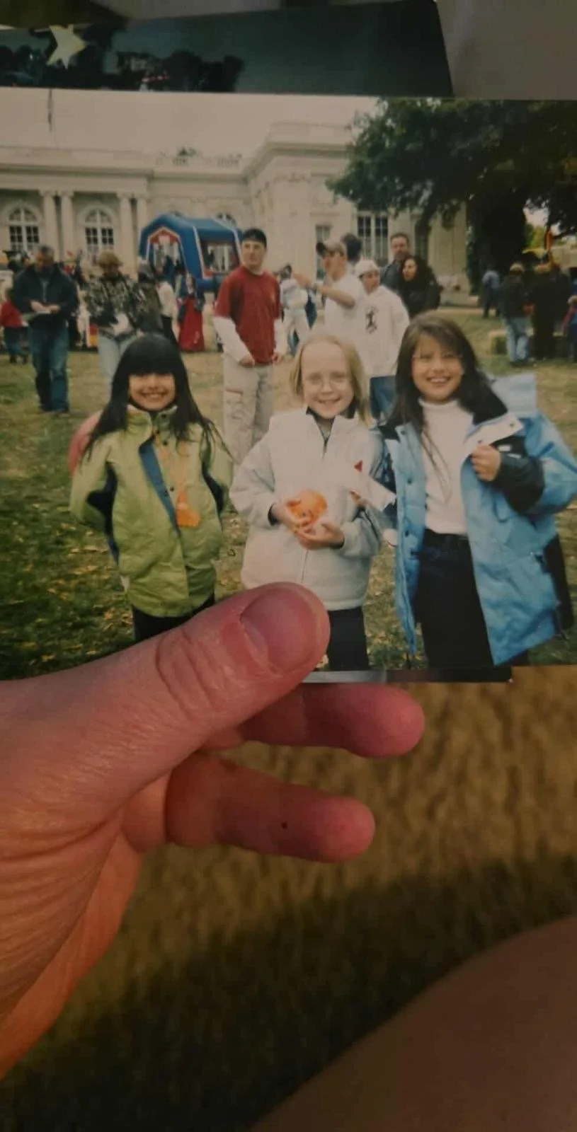 A hand holding a photograph of three smiling kids, two girls and one boy, at an outdoor event in front of a historic building, with a crowd and trees in the background.