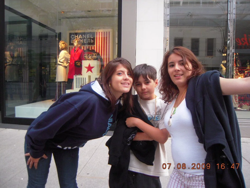 Three young women and a young boy taking a selfie in front of a designer store window display featuring mannequins dressed in fashionable clothing.