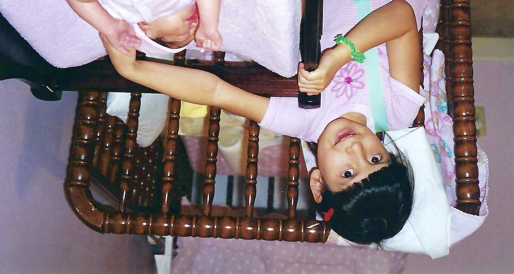 A young girl lying upside down on a bed, smiling at the camera, holding a phone with her right hand and touching another child's head with her left hand, inside a wooden bed frame.