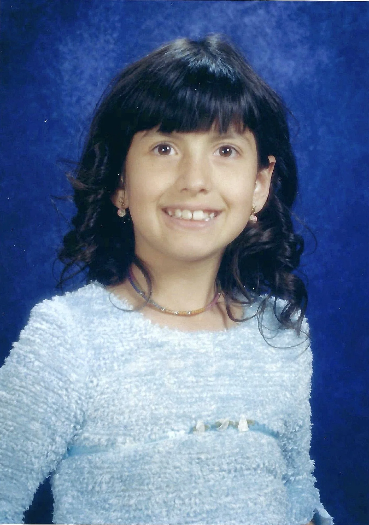 Young girl with black hair and a gap-tooth smile, wearing earrings, a necklace, and a white textured sweater, sitting against a blue backdrop.