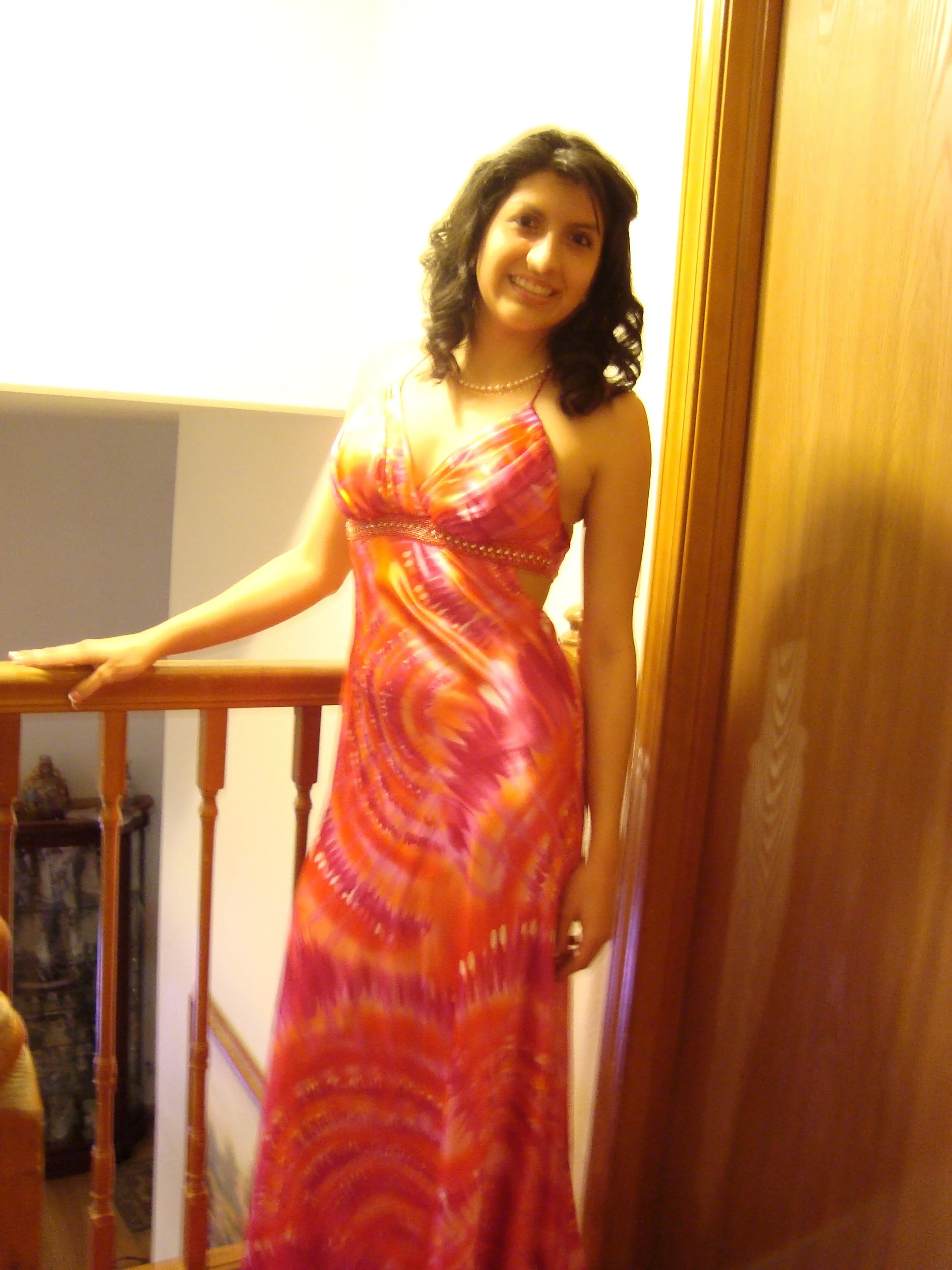 A woman in a colorful pink and orange dress with a pearl necklace standing indoors near a wooden door and railing, smiling at the camera.