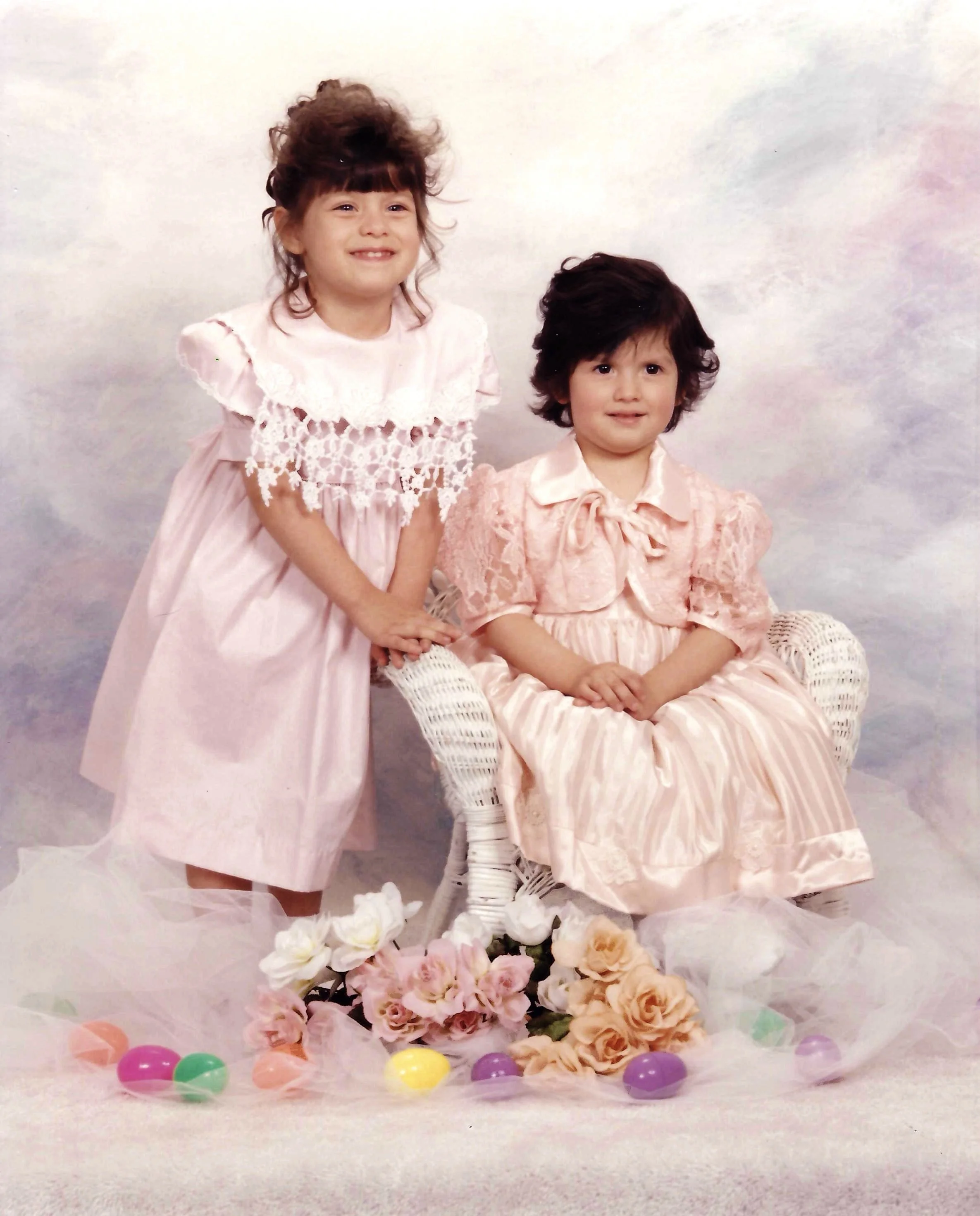 Two young children with vintage clothing and hairstyles, surrounded by flowers and Easter eggs, in front of a pastel-colored background.