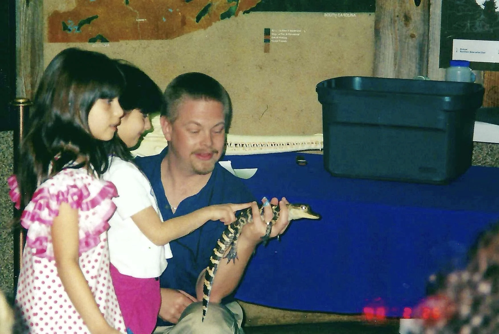 A man sitting at a table holding a small alligator while two young girls observe closely.